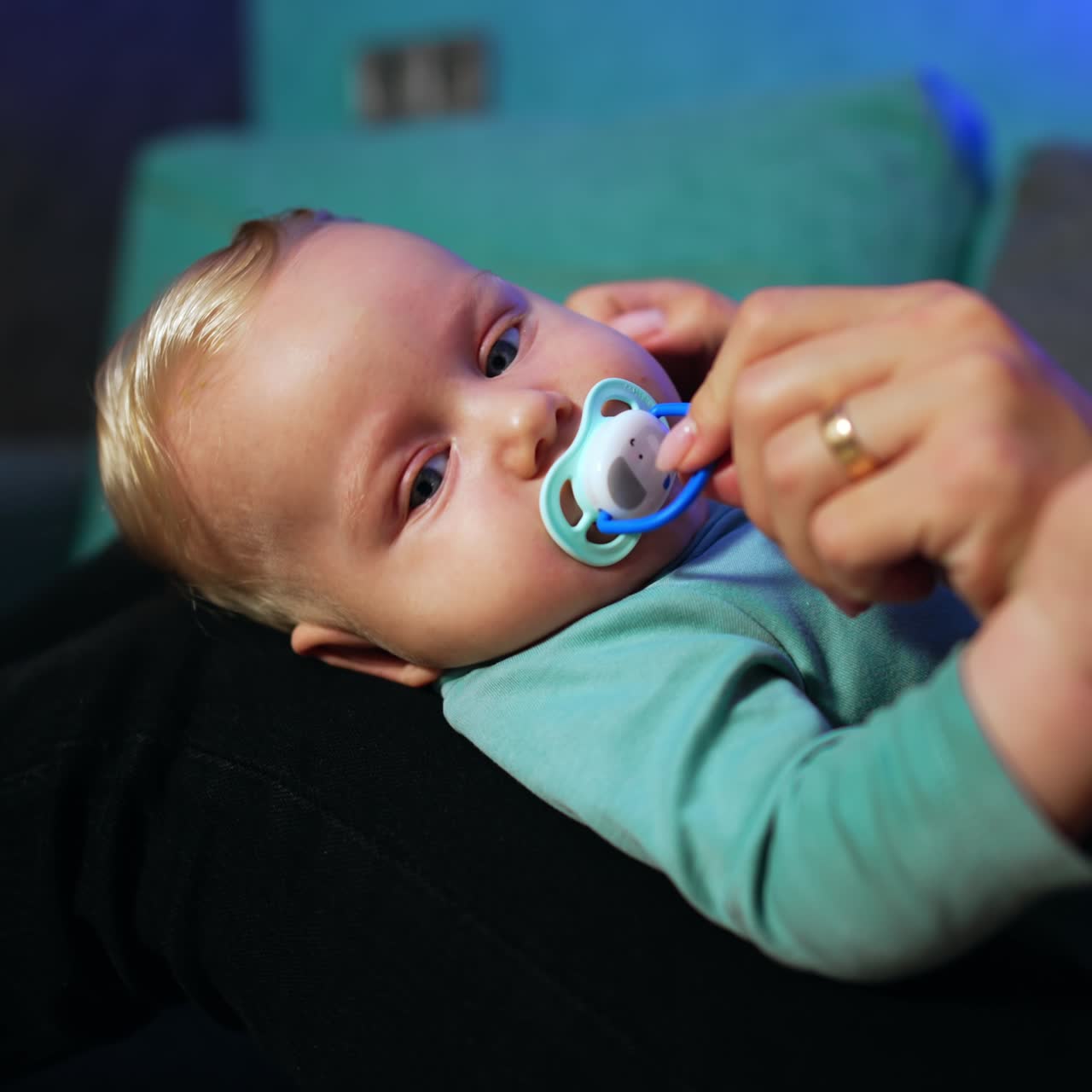 Lovely child with fair hair lies on mom's laps. Mother takes a pacifier out of kid's mouth and waves it in front of child's face. Close up