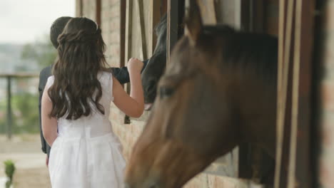 a boy in an elegant suit and a girl in a white dress tenderly caress a horse from a stable window.