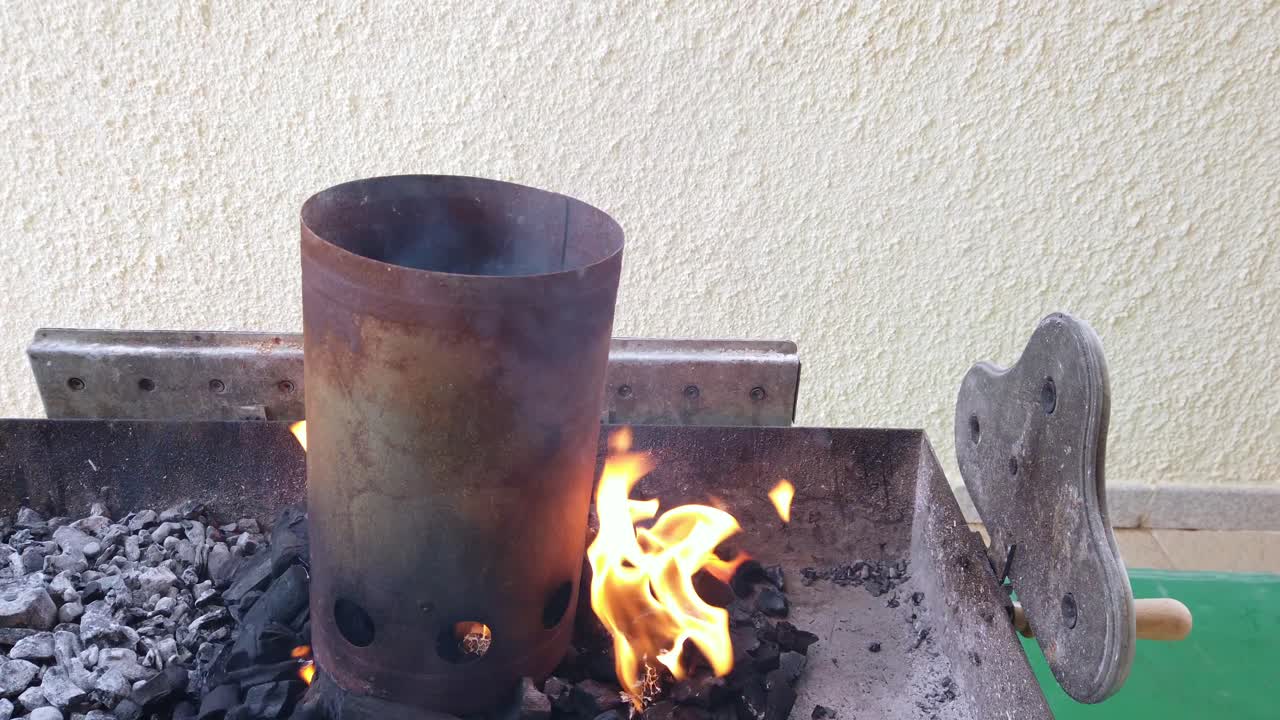 Zapalování dřevěného uhlí v grilovací vaně k založení grilovacího ohně. Detail plamenů vzněcujících se a šířících se po uhlí v grilu. Příprava venkovního grilování s přirozeným ohněm, žárem a kouřem.