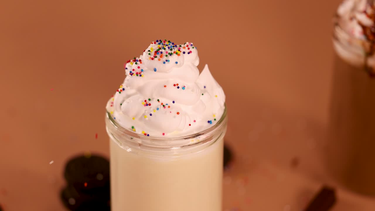 Colorful sprinkles fall onto whipped cream-topped vanilla milkshake in glass jar, brown background