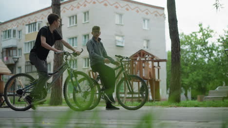 two boys sit on their bikes interacting in a park setting, one boy rests while holding his handlebars, looking towards the other boy, in the background, there is another child riding a scooter