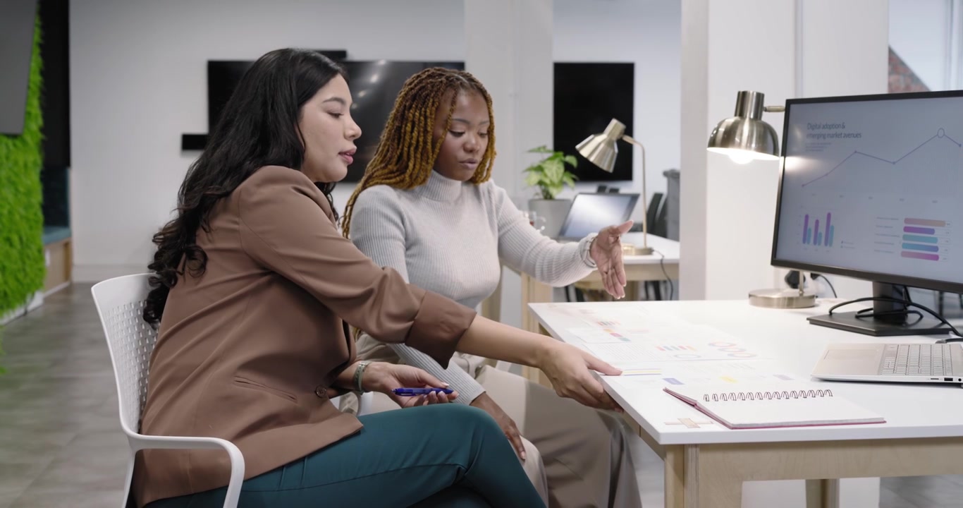 Diverse colleghe donne che esaminano grafici alla scrivania, che studiano grafici sul monitor, che indicano, che smistano documenti