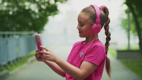 a young girl enjoys listening to music with pink headphones while using her smartphone, embodying a carefree and joyful moment in a serene outdoor setting