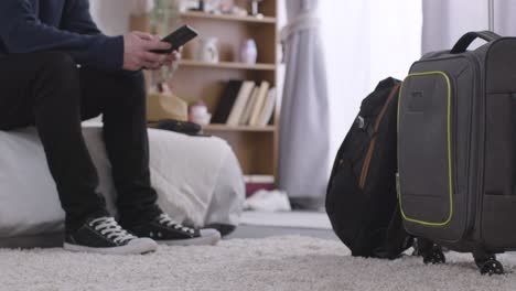 man at home with luggage waiting to leave for summer holiday checking mobile phone