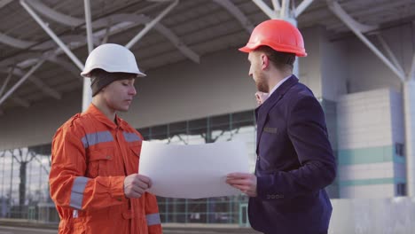 Joven-Gerente-Del-Proyecto-Con-Traje-Negro-Que-Examina-El-Objeto-De-Construcción-Con-Un-Trabajador-De-La-Construcción-Con-Uniforme-Naranja-Y-Casco.