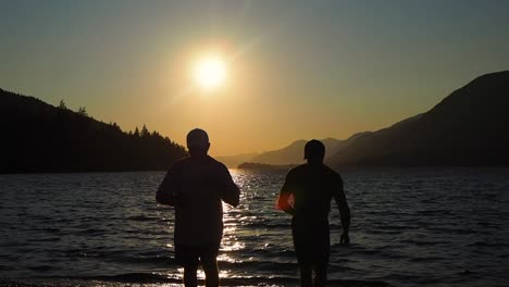 sunset on a lake with mountains in the background with two guys having a drink in silhouette