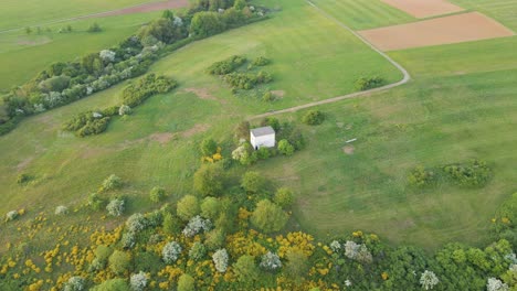 Brick-hut-sitting-at-the-end-of-a-countryside-dirt-road-surrounded-by-lush-fields-and-blossoming-bushes-and-trees