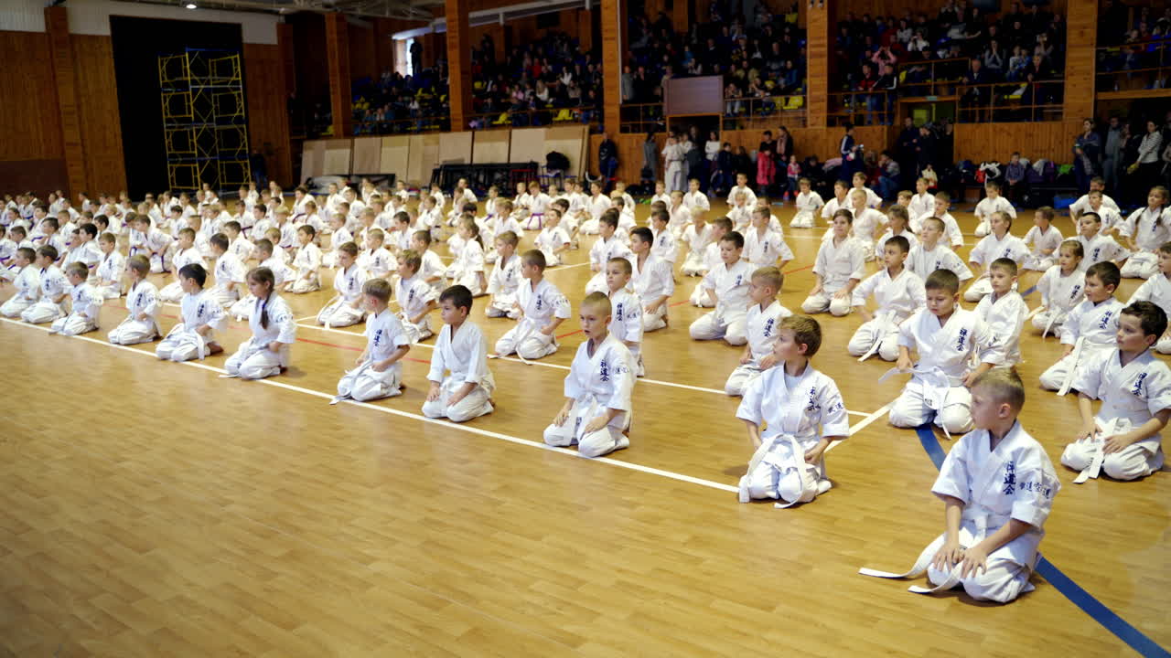 Karate trainees sitting on the floor and doing the bows. Young sportsmen stand up and stand in rows. Audience at backdrop.