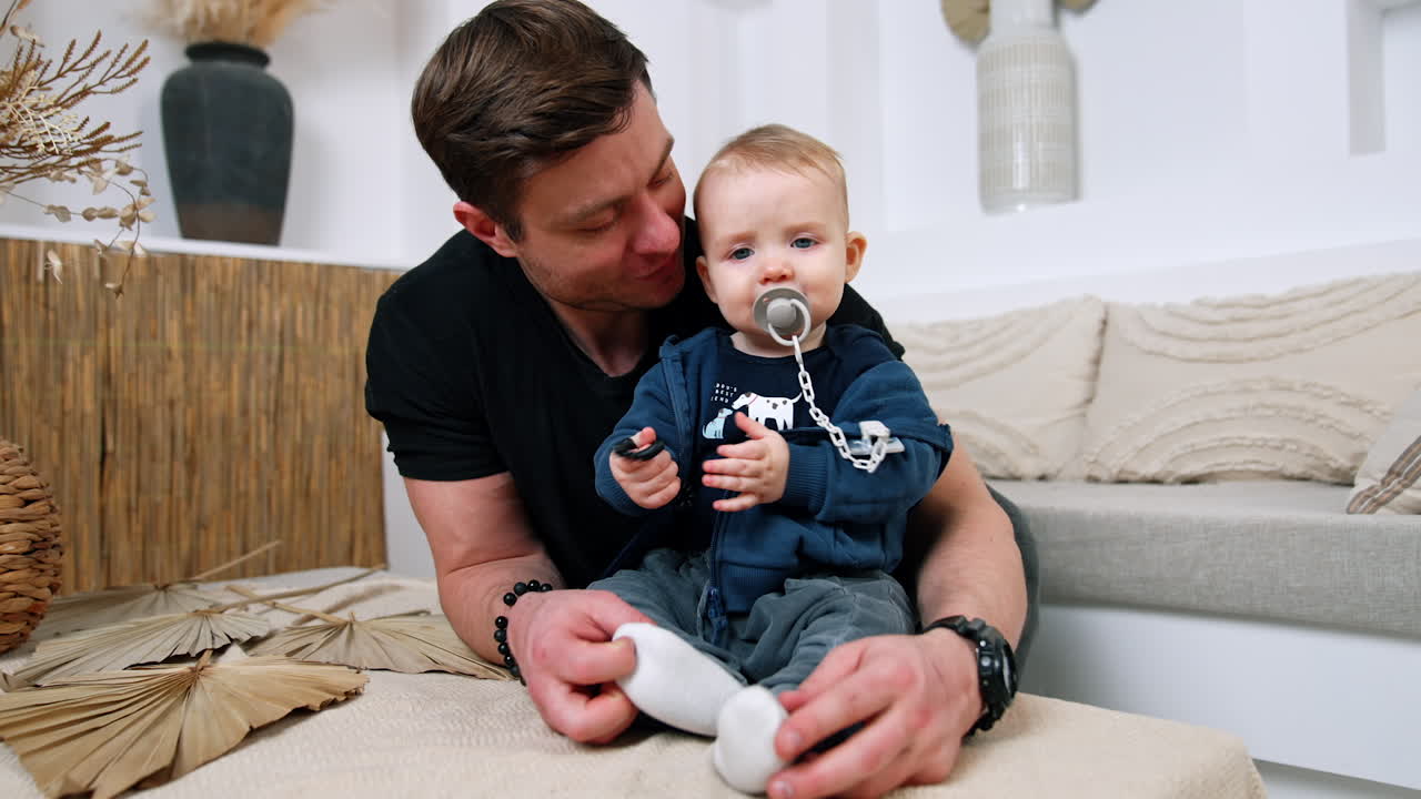 Adorable baby boy with pacifier in mouth and bracelet in hands sits on the coffee table. Loving dad embraces a child and touches his little feet.