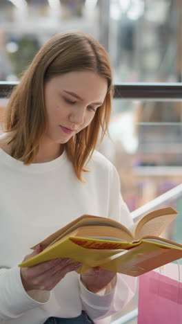 mujer joven sentada en un centro comercial moderno leyendo libro amarillo pensativo con bolsas de compras cerca, barandillas de vidrio contemporáneas y reflejos de luz suave