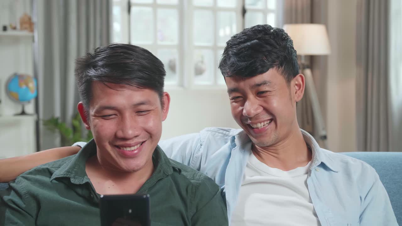 Portrait Of Asian Gentle Gay Couple Using Mobile Phone, While Sitting On A Couch In Cozy Stylish Apartment. Adult Boyfriends Online Shopping On Internet, Watching Funny Videos On Streaming Service