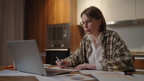 portrait of a woman with glasses working remotely in a home office at a desk with a laptop and notes data on a graph business economist analyst