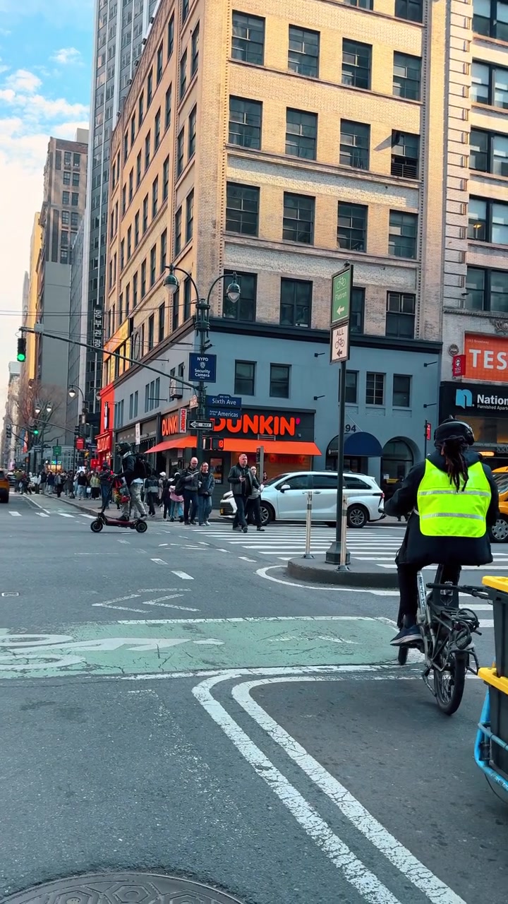 New York, USA, 22 March 2026: Pedestrians crossing city street intersection near coffee shop facade. Busy urban scene at 6th Avenue New York with people walking and delivery cart