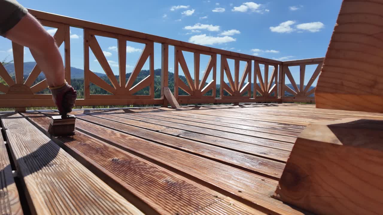 Hand in glove oils wooden terrace boards with brush. Decorative railing, serene sky, clouds, and mountain horizon evoke home improvement and outdoor maintenance themes