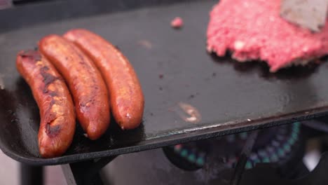 Wurst-Verbindet-Fleischgrillen-Auf-Gusseisernem-Bratpfannengrill-Mit-Hamburger-Rindfleischpasteten-Im-Hintergrund-Und-Spachtel,-Der-Lebensmittel-Mit-Propangasflamme-Unter-Der-Pfanne-Bewegt