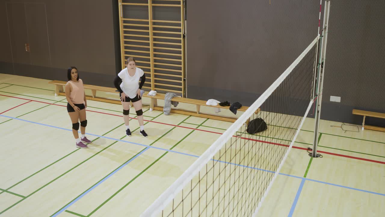 Různorodé spoluhráčky ve sportovním oblečení se pohybují u sítě v tělocvičně a reagují na přilétající míč.
