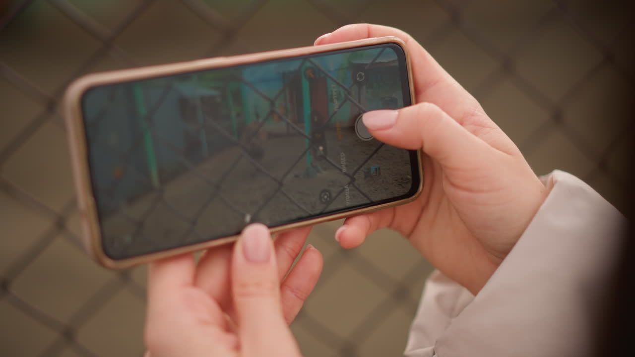 Mani che tengono uno smartphone mentre riprendono un parco giochi attraverso una recinzione metallica, dita che toccano lo schermo con profondità di campo ridotta, motivo sfocato della recinzione in primo piano, altalene vuote e graffiti visibili