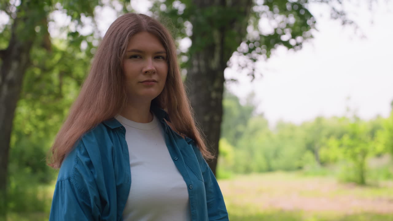 Elegant chic removes sunglasses while posing gracefully in lush green park, wearing blue shirt and white top, surrounded by vibrant foliage, expressing natural poise