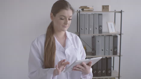 doctora pelirroja con bata blanca usando una tableta y mirando la cámara