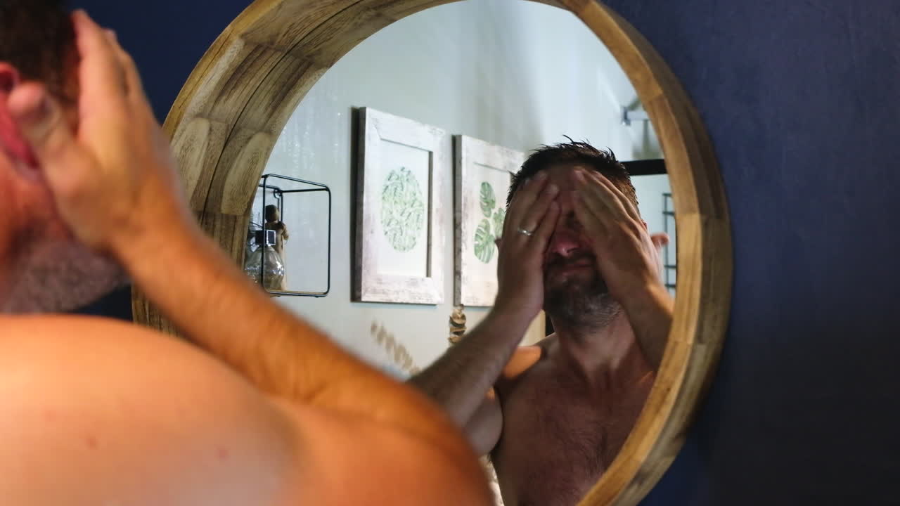 젊은 남자는 화장실에서 둥근 거울을 사용하여 얼굴 크림을 적용하고 어깨를 바라보고 있습니다.