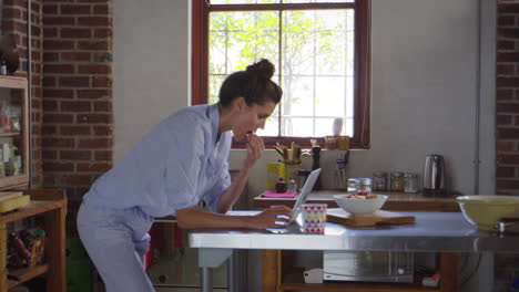 Young-woman-in-pyjamas-using-laptop-in-kitchen,-shot-on-R3D