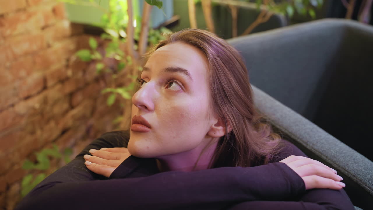 Lady with blond hair leans on cushion and looks upward in cozy indoor setting with exposed brick wall and surrounding greenery, soft light creating calm atmosphere as leaves hang near her face