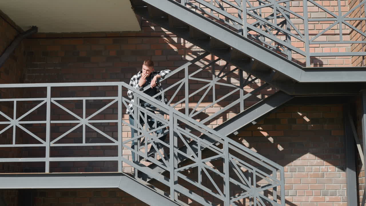 Young man in black and white checkered shirt stands on metal balcony staircase smoking cigarette with relaxed posture against brick wall as diagonal sunlight casts shadows on patterned iron railing