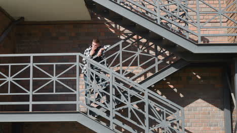 young man in black and white checkered shirt stands on metal balcony staircase smoking cigarette with relaxed posture against brick wall as diagonal sunlight casts shadows on patterned iron railing