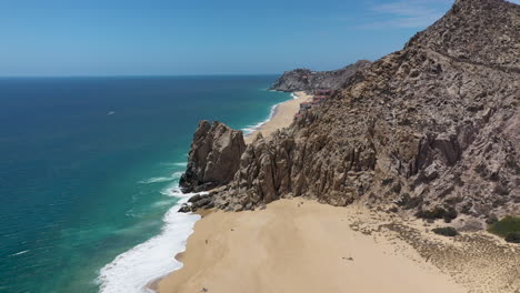 Drone-shot-of-sea-cliffs-and-beaches,-revealing-resorts-on-Playa-El-Médano-in-Cabo-San-Lucas-Mexico,-wide-and-rotating