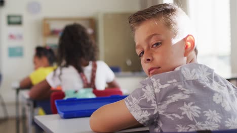 retrato de un niño de escuela caucásico feliz sentado en el aula, tomando notas, mirando a la cámara