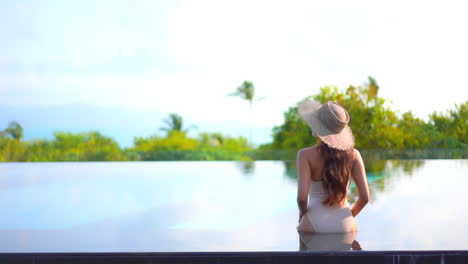 vista trasera de la mujer sentada al borde de la piscina infinita en un hotel exótico en bali usando monokini y sombrero para el sol