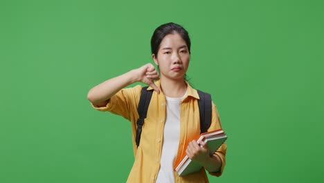 녹색 스크린 배경 스튜디오에 서 있는 동안 머리를 흔들고 엄지손가락을 아래로 표시하는 일부 책과 배을 가진 아시아 여성 학생