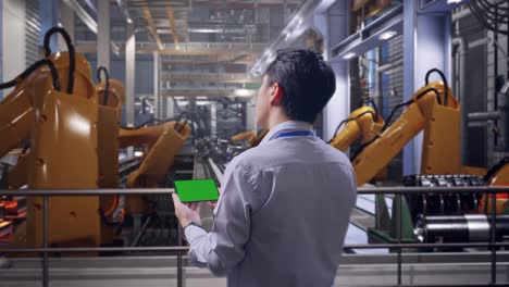 back view of an asian male professional worker standing with industrial robotic arms working, checking on smartphone with green screen and look at the factory once