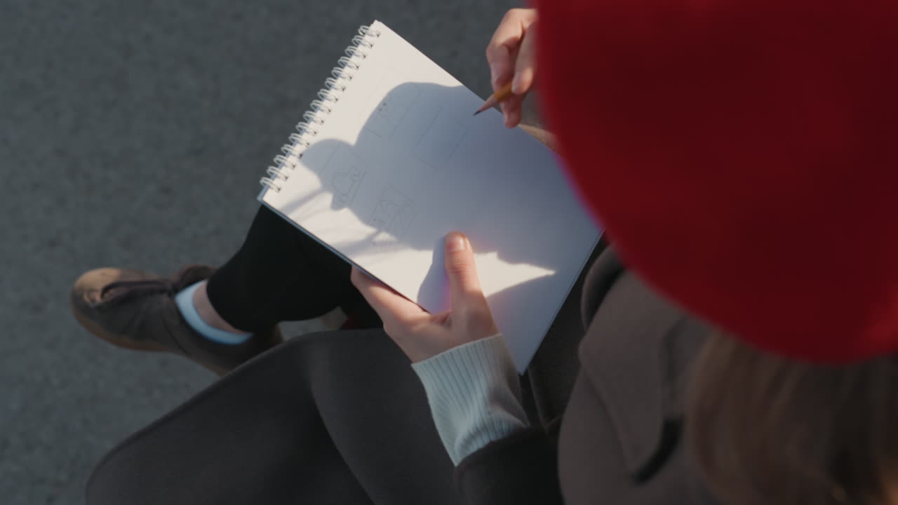 Notebook scene with focused student. Overhead perspective capturing student jotting ideas in notebook. Scene showing student with pencil and red beret writing in notebook on sunny pavement