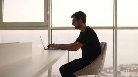 Side-view-of-business-man-using-laptop-computer-at-remote-workplace-in-slow-motion.-Freelancer-working-on-computer-at-home.-Focused-man-looking-laptop-at-big-house-with-panoramic-windows