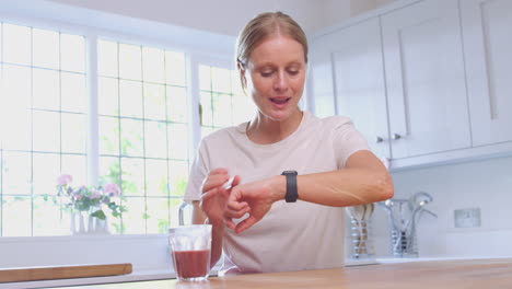 Reife-Frau-In-Fitnesskleidung-Mit-Gesundheitsgetränk-Zu-Hause-In-Der-Küche-überprüft-Die-Aktivität-Auf-Einer-Smartwatch