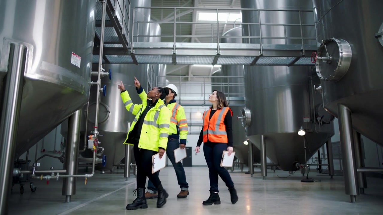 Entering plant, three workers in hi-vis inspecting, pointing at stainless tanks, piping, clipboards