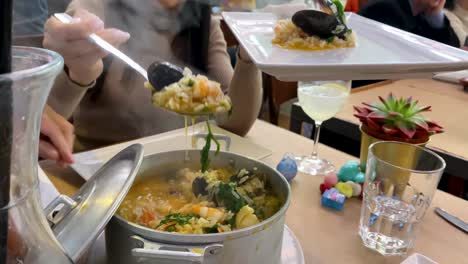 a pretty woman serves hot seafood rice from a pot onto a white plate at the cascais market