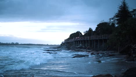 Waves-crash-from-the-ocean-into-the-shore-underneath-a-long,-raised,-wooden-boardwalk-that-sits-looking-over-the-ocean-as-the-sunset-spills-a-blue-tint-into-the-sky-filled-with-fluffy-clouds