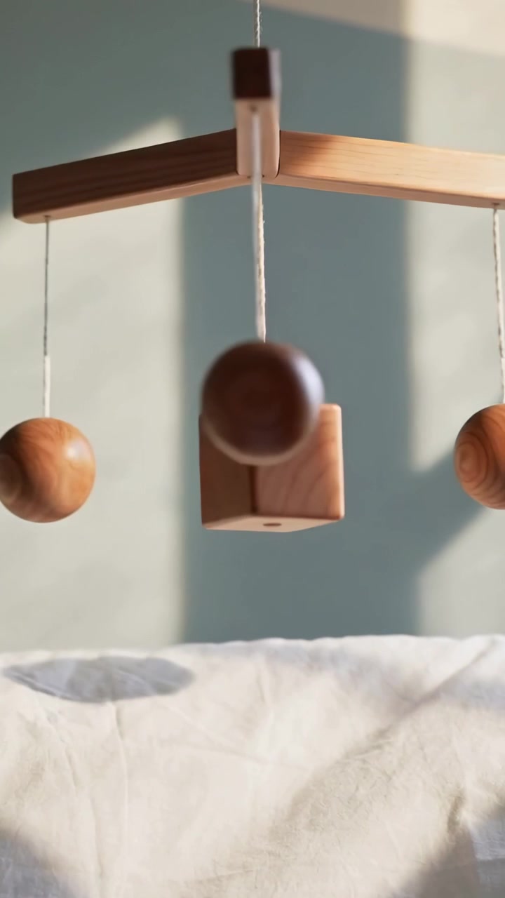 Vertical video: Swaying mobile with cube and beads above quilt near window, sunlight moving on wall