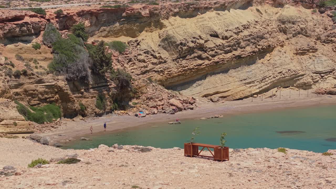 Un filmato aereo mostra la spiaggia di Agios Thodoros a Karpathos da una panchina all'ingresso, mettendo in evidenza le limpide acque turchesi, il terreno aspro e la bellezza selvaggia dell'isola.