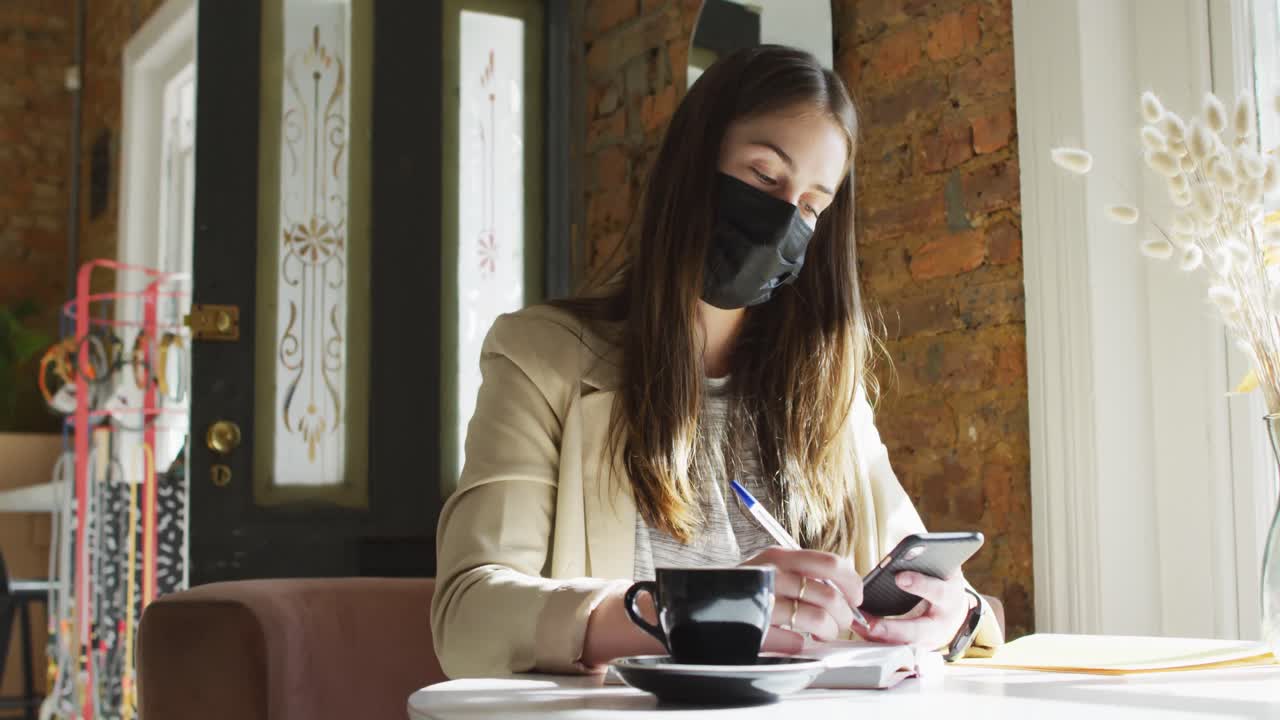 Clienta caucásica con mascarilla, sentada a la mesa, usando un smartphone.