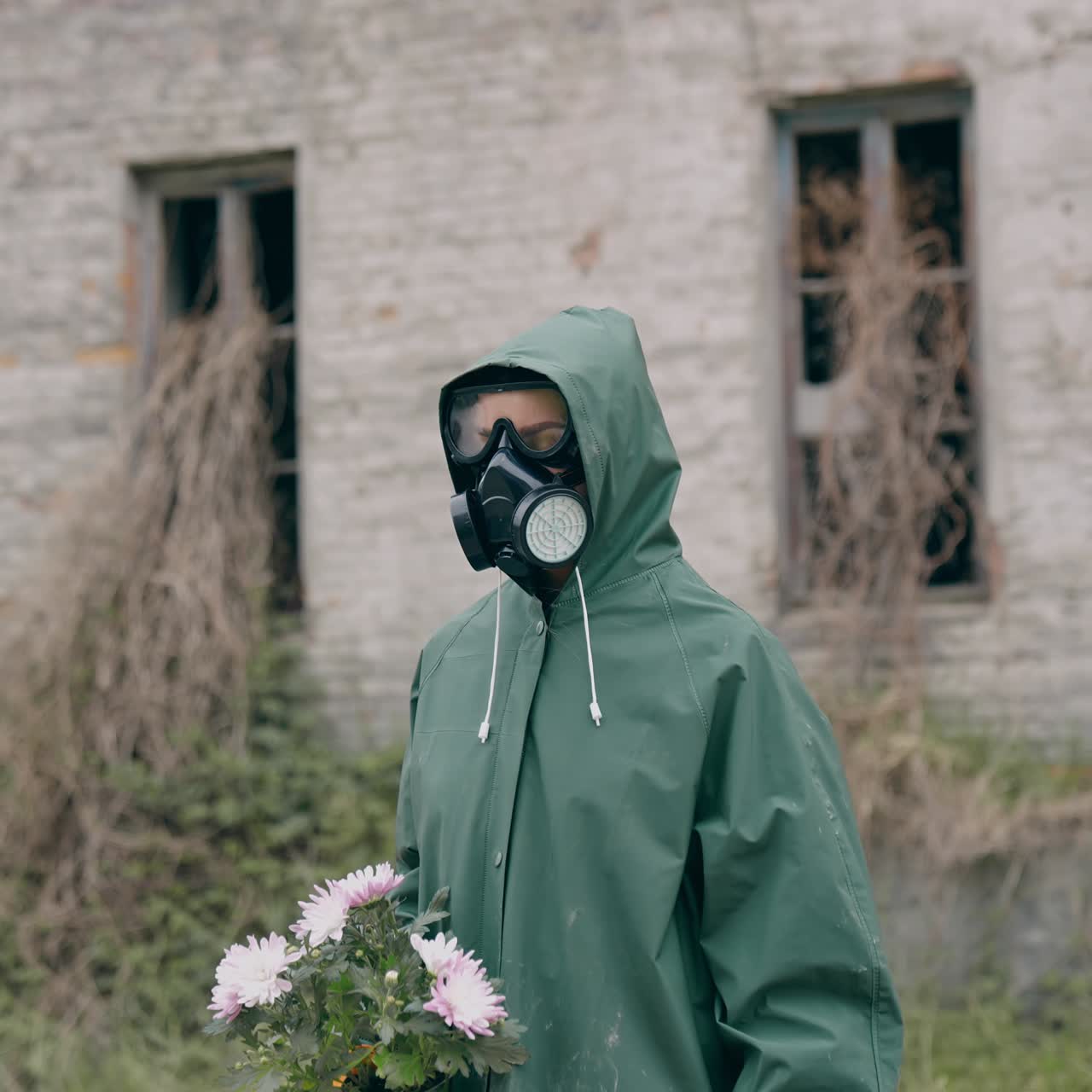 Survivor holding flowers on ruined building background. Person in chemical safety suit and respirator standing with flowers in hands in abandoned place. Chemical attack