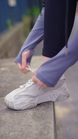 Vertical-Video-Close-Up-Of-Woman-Tying-Laces-On-Training-Shoe-Before-Exercising-Running-Along-Urban-Street