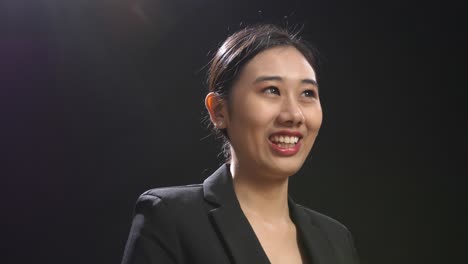 side view of smiling asian speaker woman in business suit showing thumbs up gesture while standing in the black screen studio