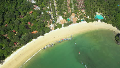 Toma-Aérea-De-Arriba-Hacia-Abajo-De-La-Playa-De-Arena-Con-Botes-De-Estacionamiento-Y-Pueblo-En-El-Paisaje-En-Un-Día-Soleado---Playa-De-Railay,-Krabi,-Tailandia