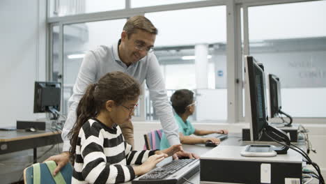 chica enfocada en anteojos sentada y haciendo tareas en la computadora mientras la maestra la mira