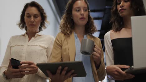 Diverse-female-colleagues-in-discussion-using-laptop-and-tablet-in-office-corridor,-slow-motion