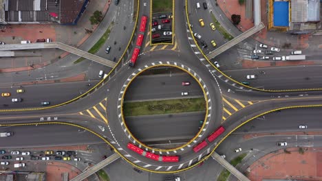 a bird's eye view shot with a 4k drone descending on a round point intersection with red articulated buses, taxis and other vehicles turning from third street to 30th avenue in bogotá, colombia