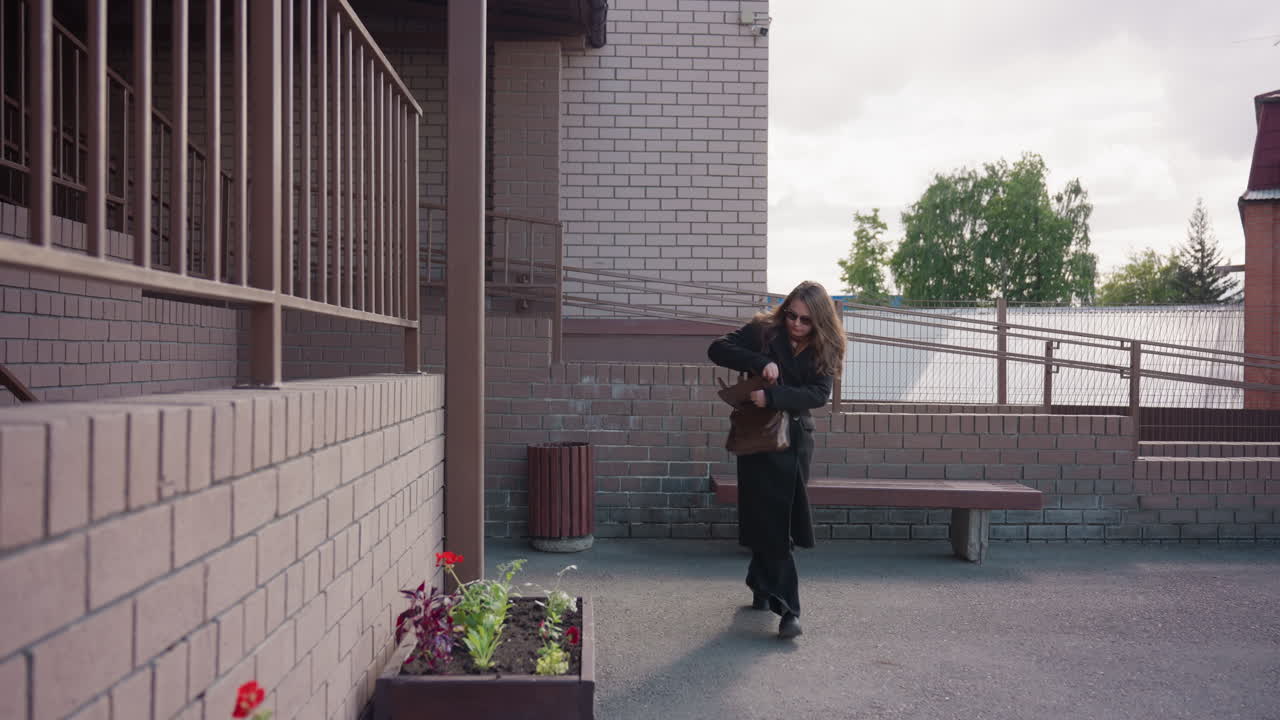 검은색 코트를 입은 비즈니스 여성이 벽돌 안뜰을 가로질러 서둘러 걸어가면서 갈색 가방 안을 확인하고, 벤치, 화분, 나무가 도시 입구를 장식하고, 목적 있는 움직임, 솔직한 거리의 순간을 보여줘요.